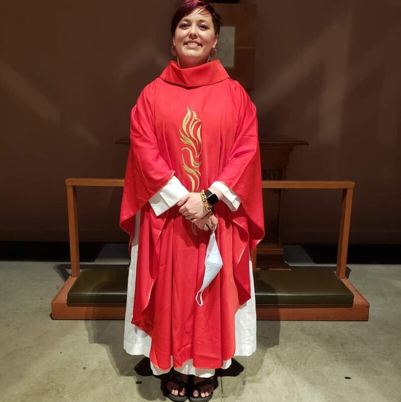 Image of a young woman wearing Pentecost clergy attire, standing on a light colored floor with a dark (wood) background