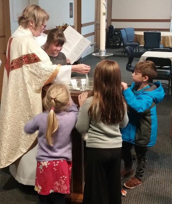 image of a female priest standing over a baptismal font with three children surrounding the wooden font