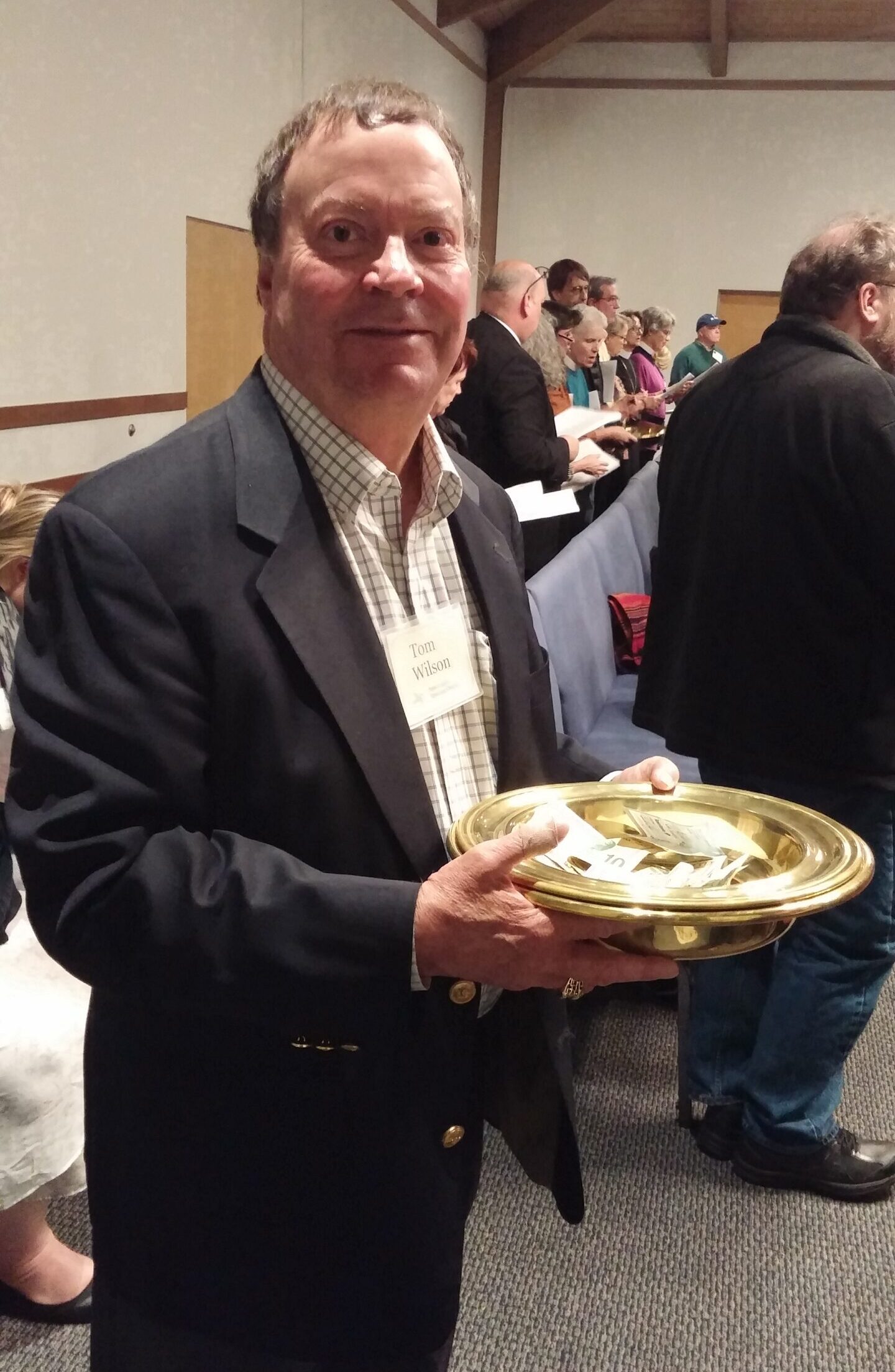 image of a middle-aged white man in a black jacket holding a brass offering plate.