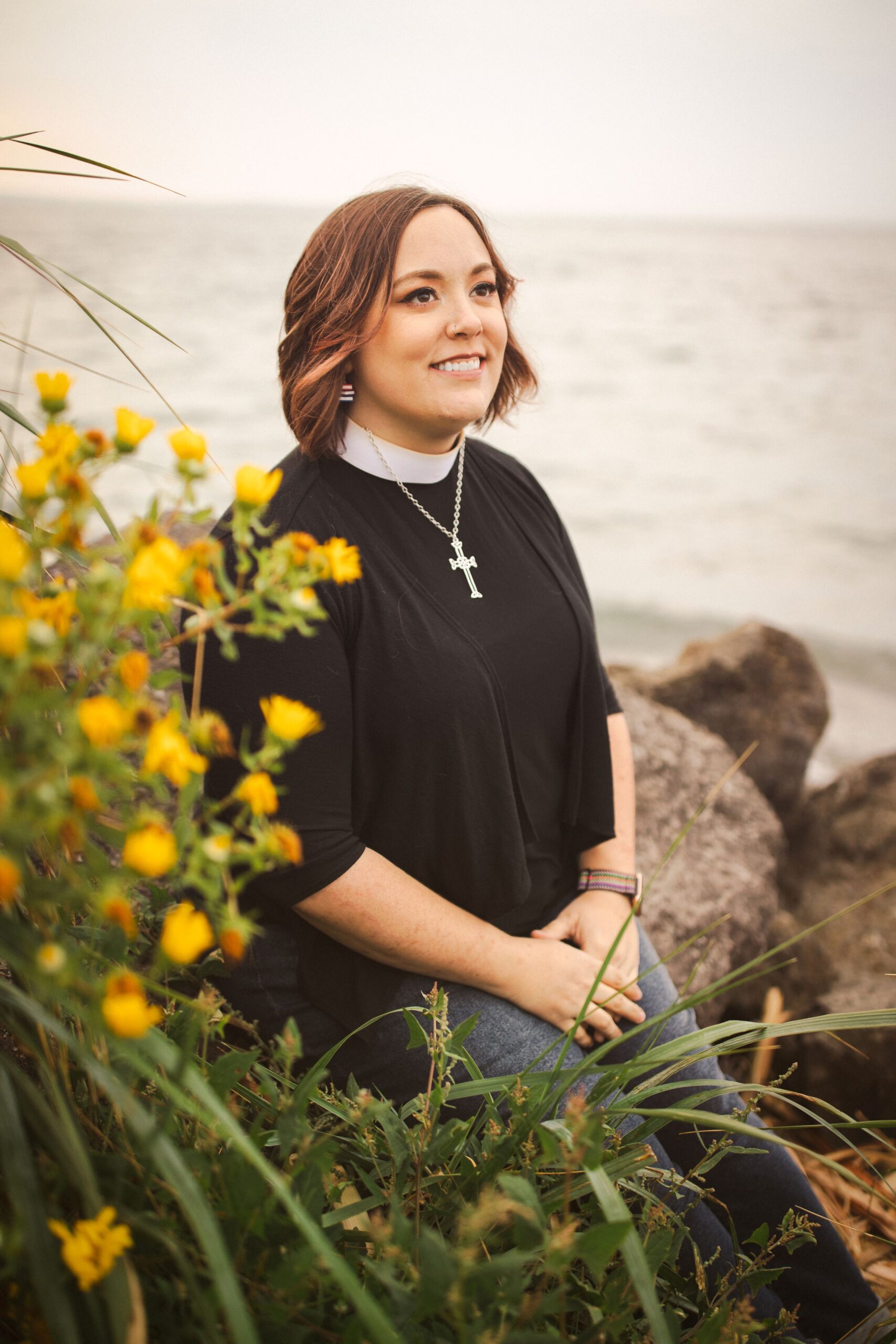 image of a young woman wearing a black shirt and clergy collar with cross necklace, partially seated, a spray of small yellow flowers in the foreground, boulders and water in the background.