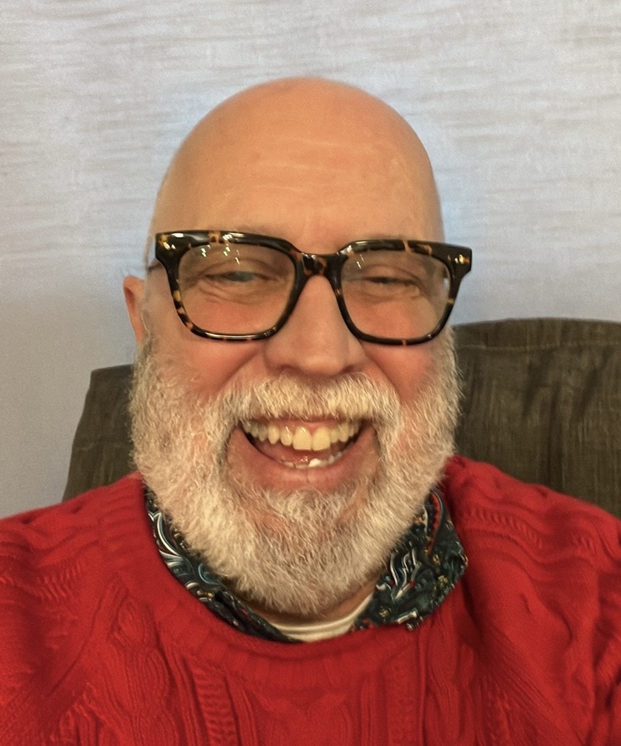 headshot with a man, smiling, bearded and with a shaved head, wearing thick frame glasses and a red sweater.
