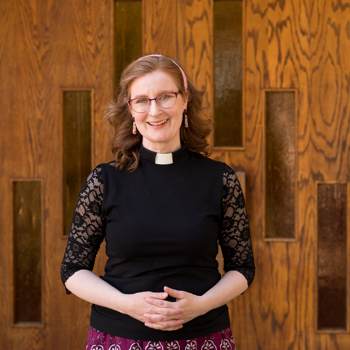 image of a woman priest wearing black shirt with lace sleeves and clergy collar standing in front of wooden door