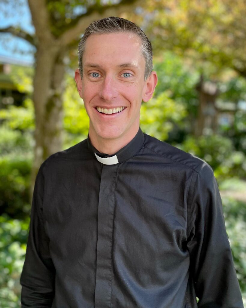 a man with short hair wearing a clergy collar and black shirt, smiling into the camera