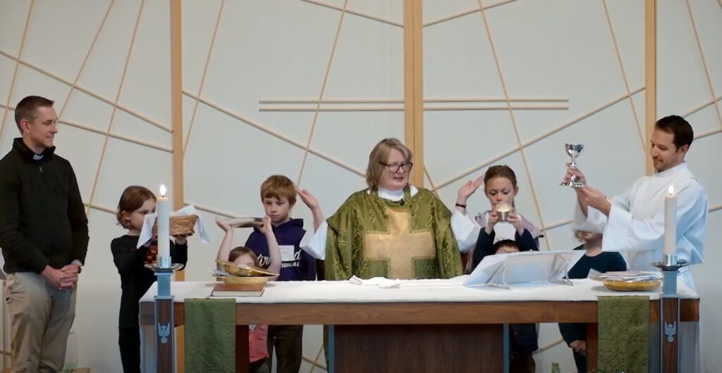 a group of kids standing behind an altar with a priest in the center and two adults on far right and far left all holding up sacramental elements for communion.