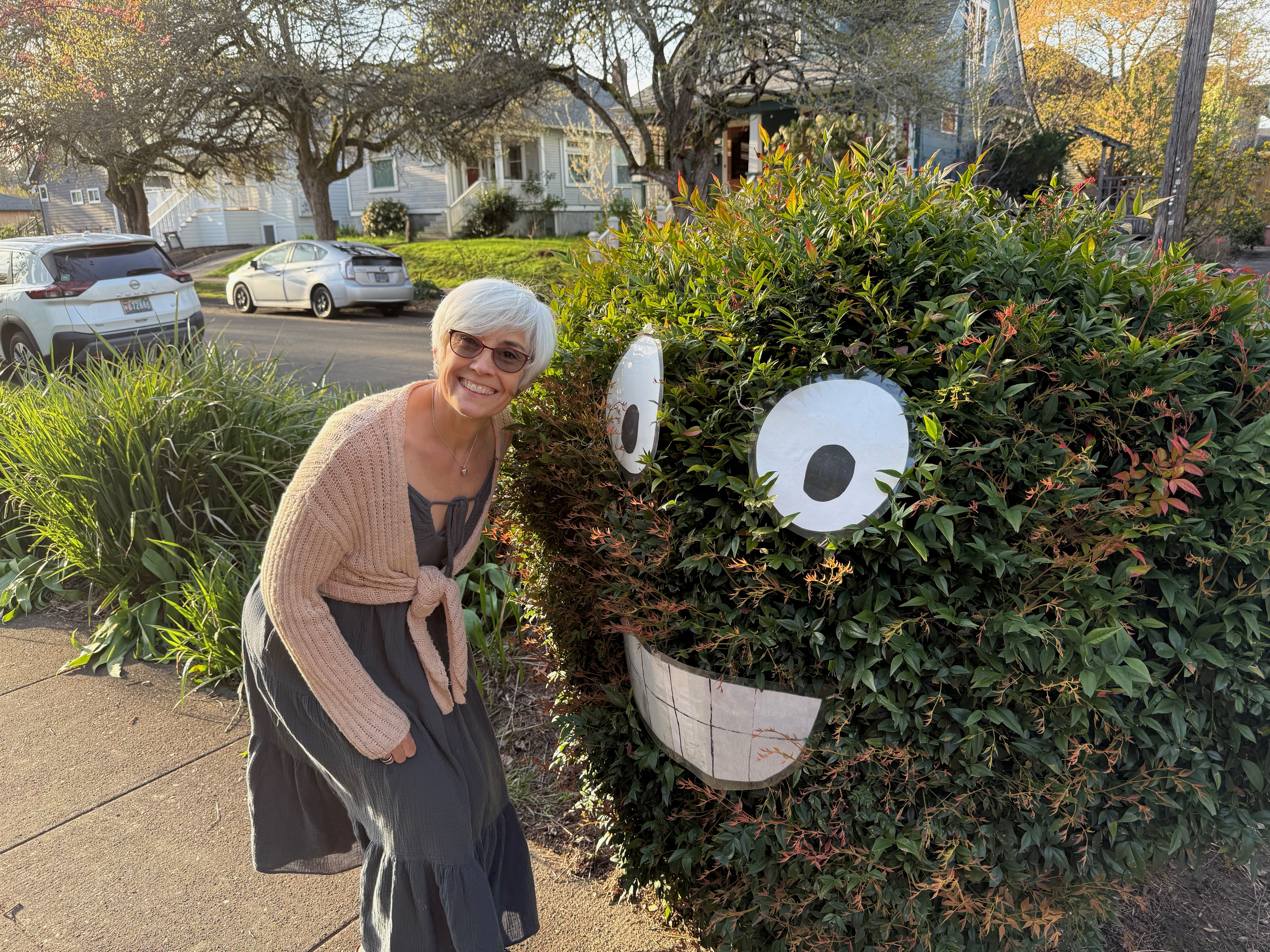 image of a woman with short grey hair and glasses smiling while crouched next to green foliage with a large smiley face.